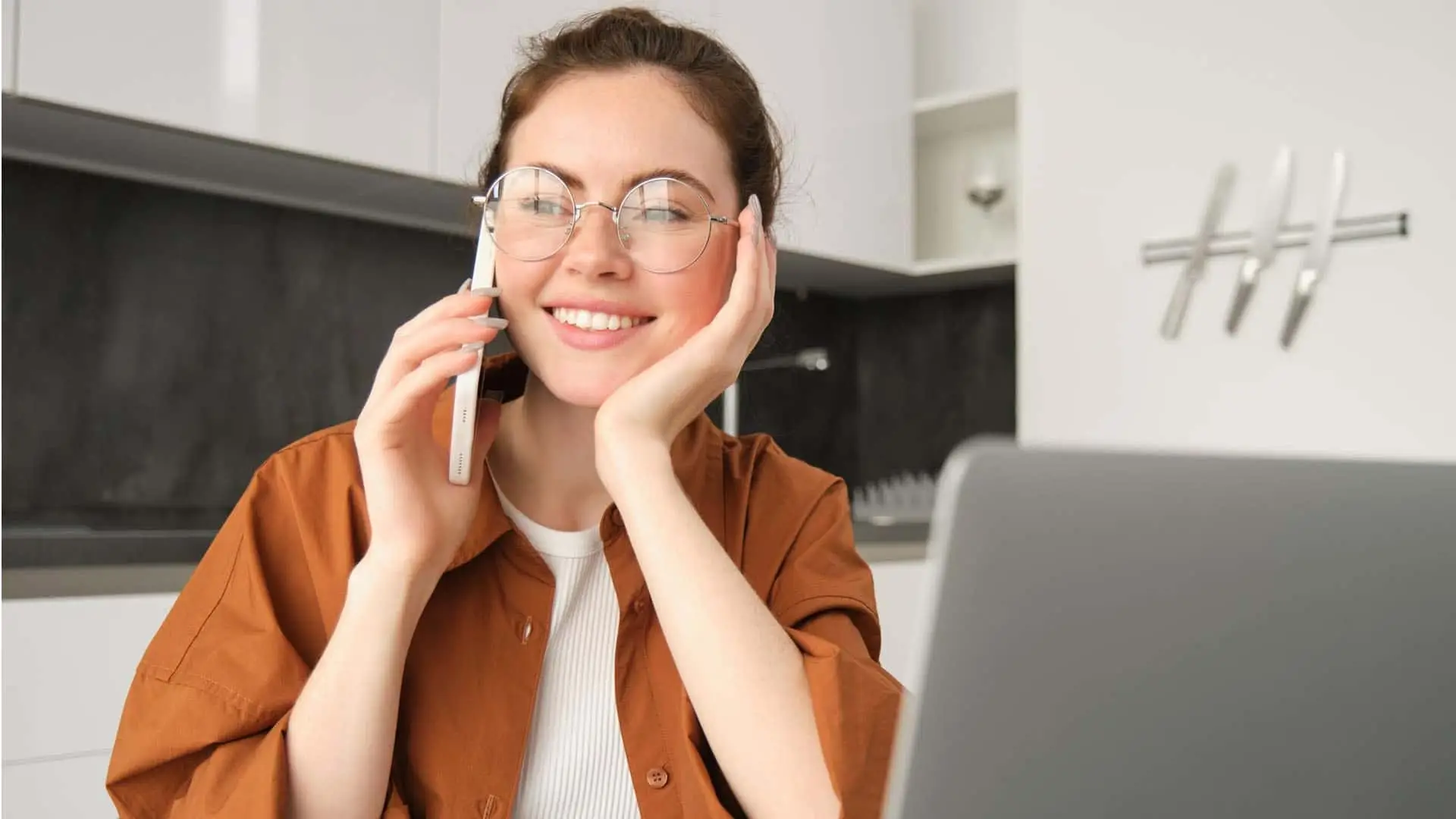 Mujer joven con gafas sonriendo mientras habla por teléfono y trabaja con su portátil en casa.