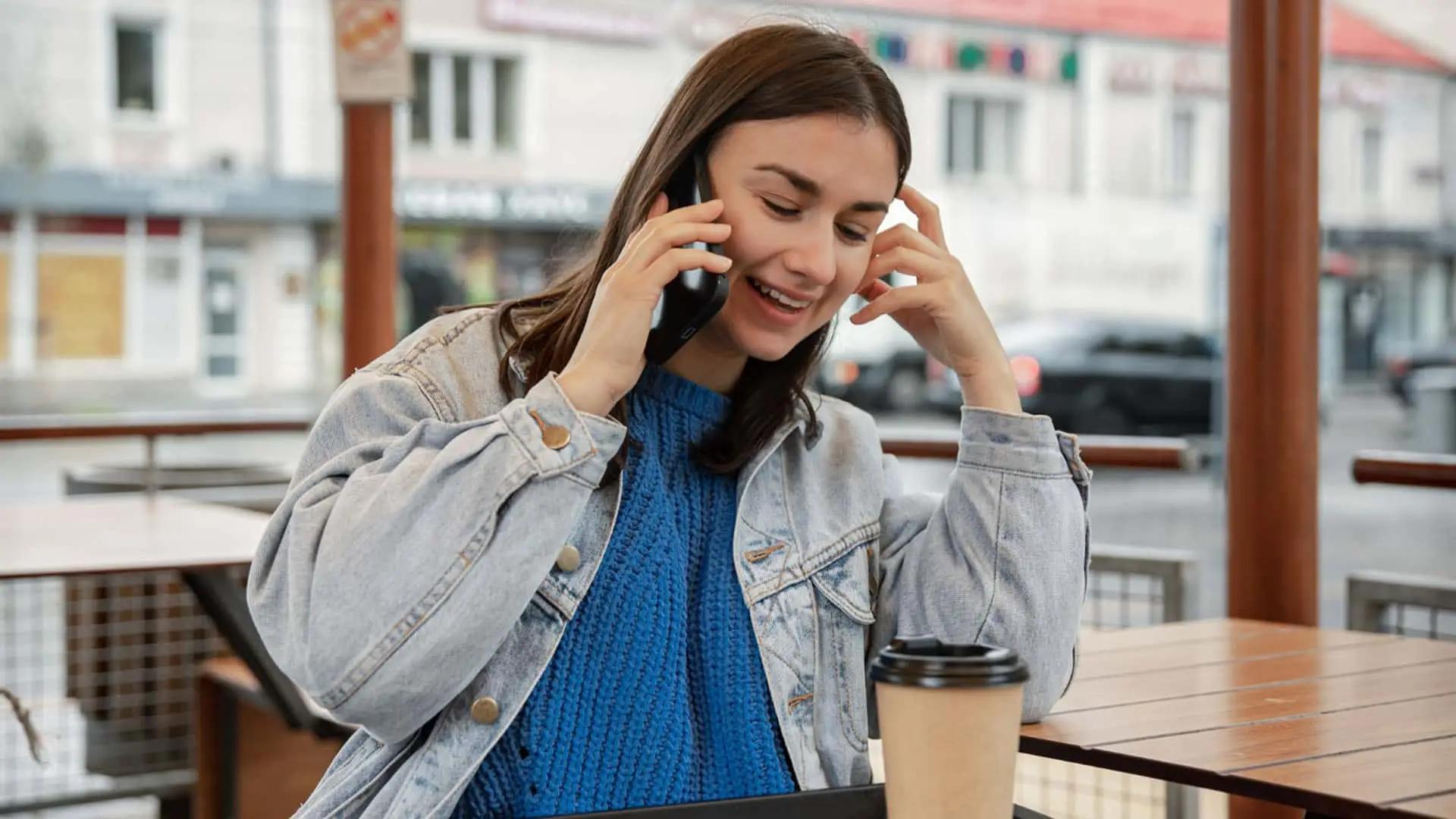 Mujer joven sonriendo mientras habla por el móvil sentada en la terraza de una cafetería.