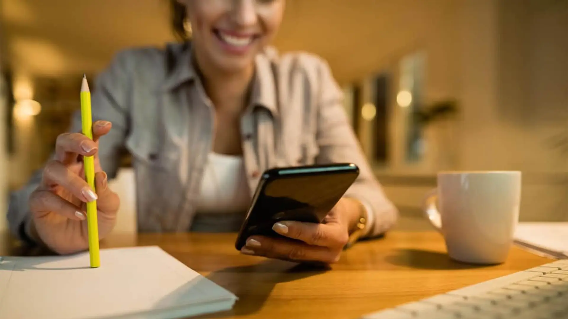 Mujer sonriente trabajando en su escritorio. Consulta el móvil mientras sostiene un lápiz sobre un cuaderno.