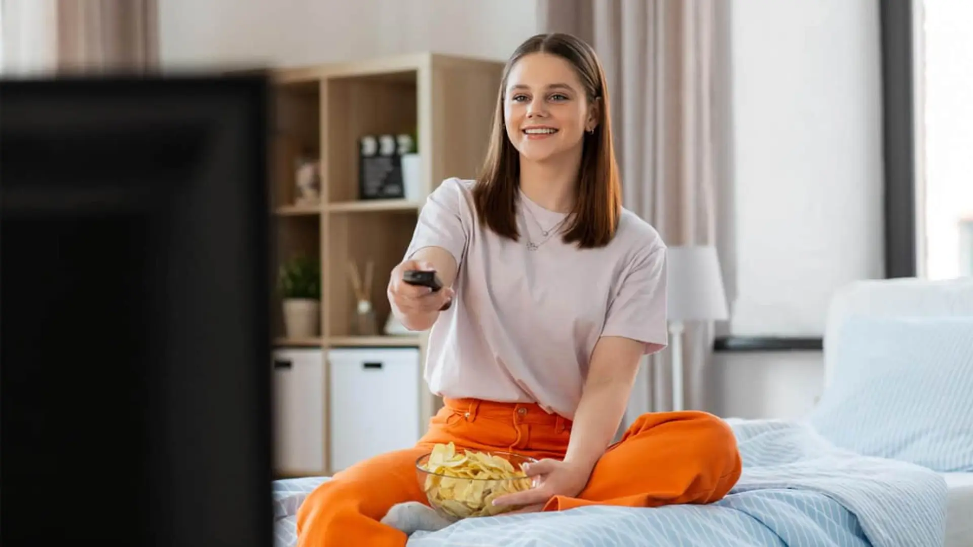 Chica joven sonriente sentada en la cama viendo la televisión con mando a distancia y un bol de patatas.