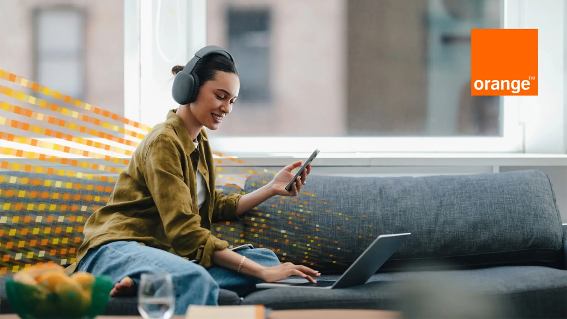 Mujer con auriculares usando portátil y smartphone en el sofá con señal digital de fondo.