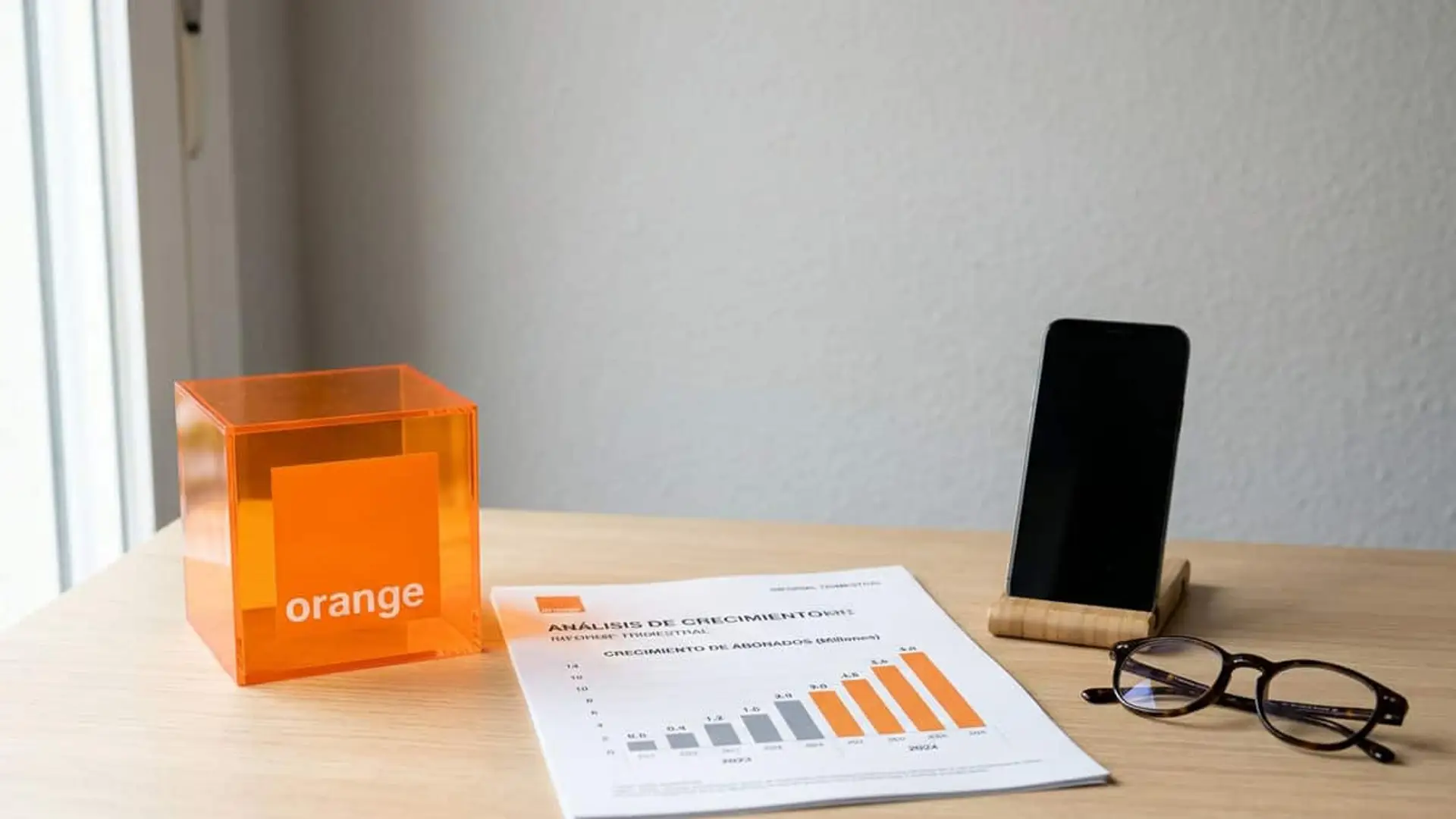 Orange España ingresa 5.673 millones hasta septiembre, un 2% más, gracias a la venta de dispositivos Logotipo de Orange junto a un informe con un gráfico de crecimiento de abonados móviles, un teléfono y unas gafas sobre una mesa.