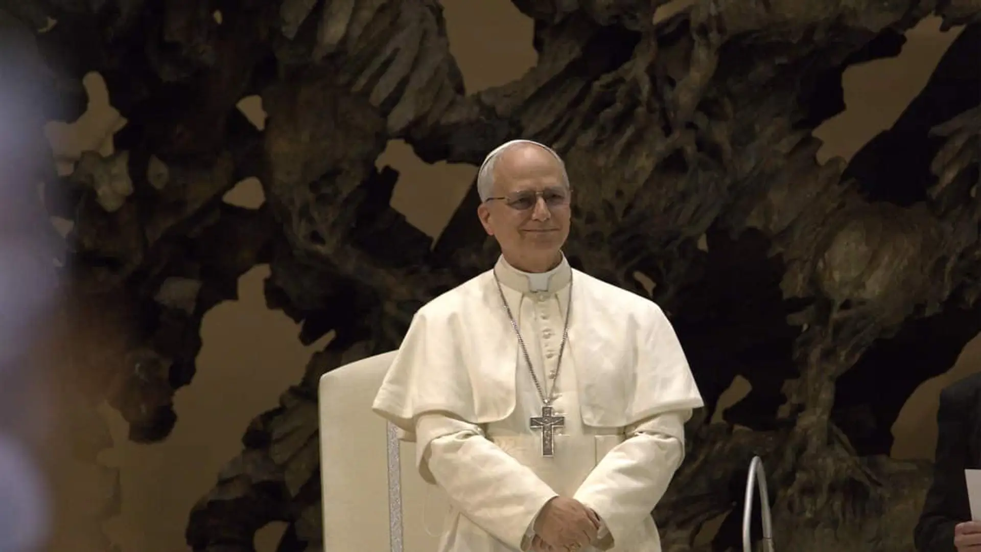 El Papa León XIV sentado con vestimenta blanca y cruz pectoral delante de un fondo oscuro