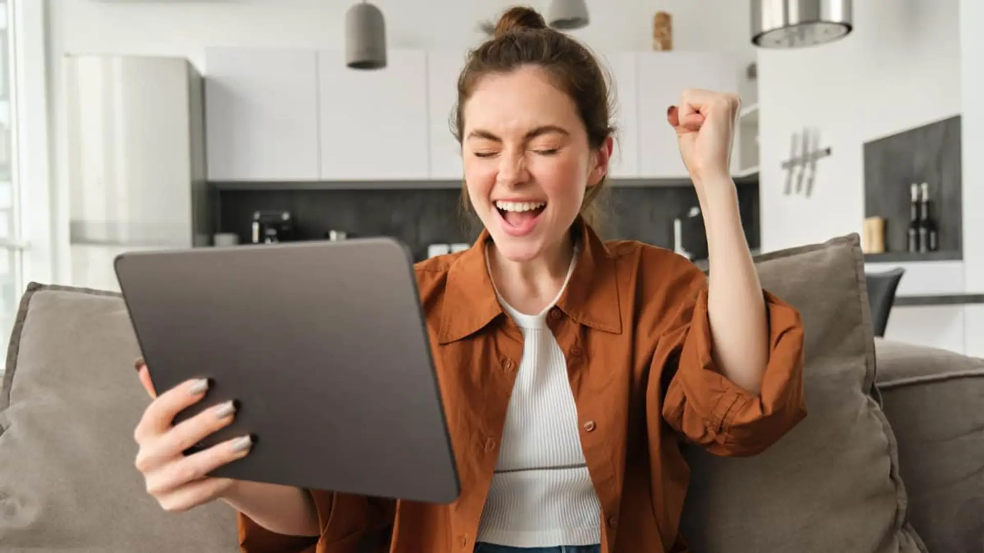 Mujer celebrando con éxito frente a su tablet en casa, representando velocidad de internet y buena conectividad.