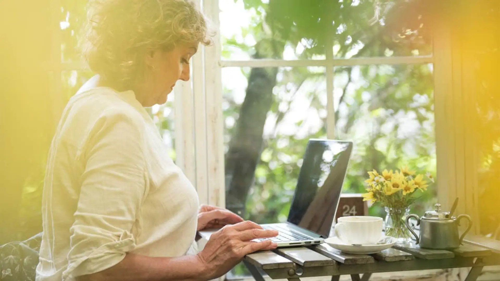Mujer adulta teletrabajando con un ordenador portátil en una terraza soleada en un ambiente relajado.