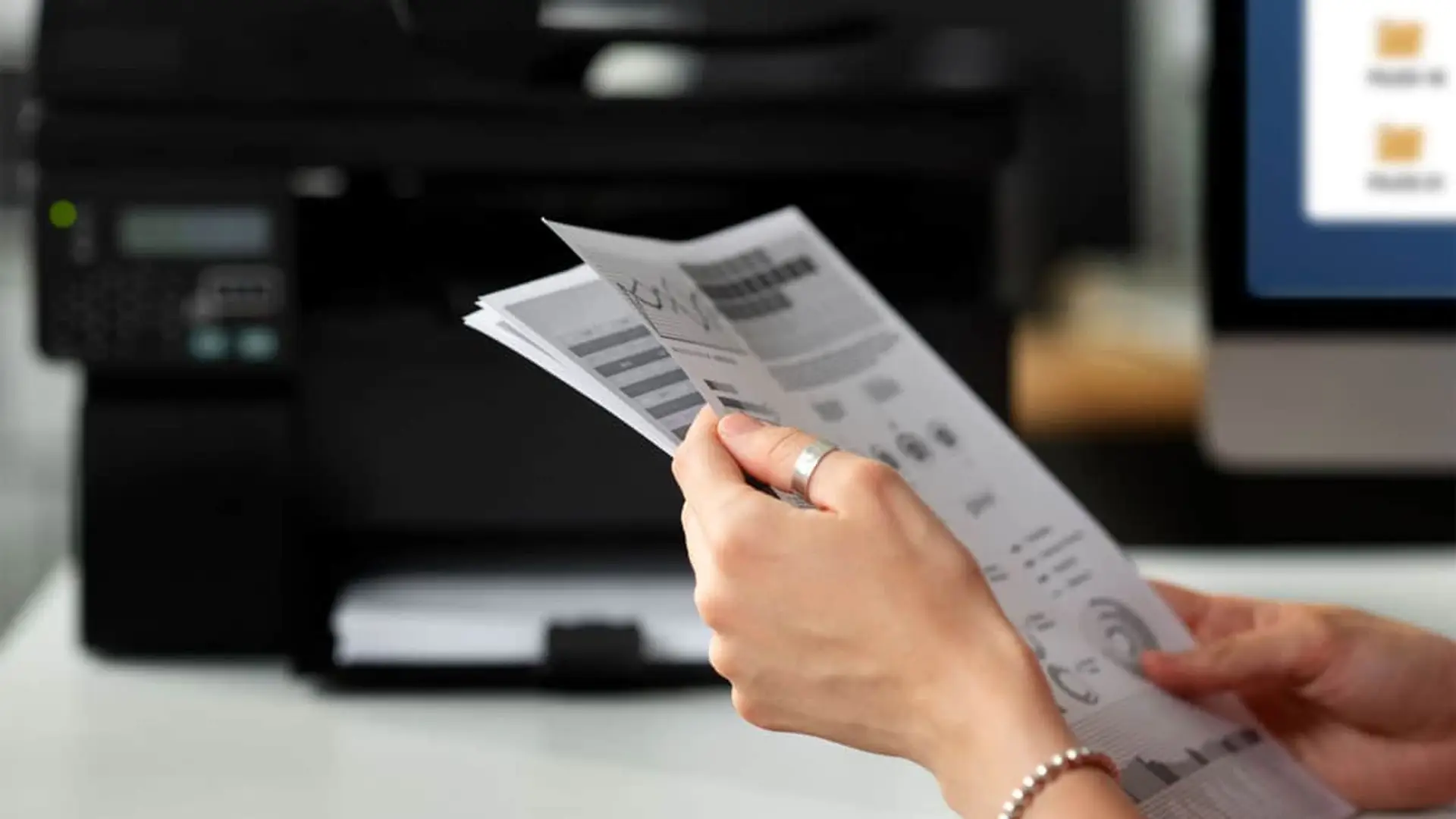 6 gastos ocultos en tu factura de móvil que estás pagando sin saberlo Primer plano de una persona revisando un documento con gráficos en una oficina, con la impresora y el ordenador al fondo.