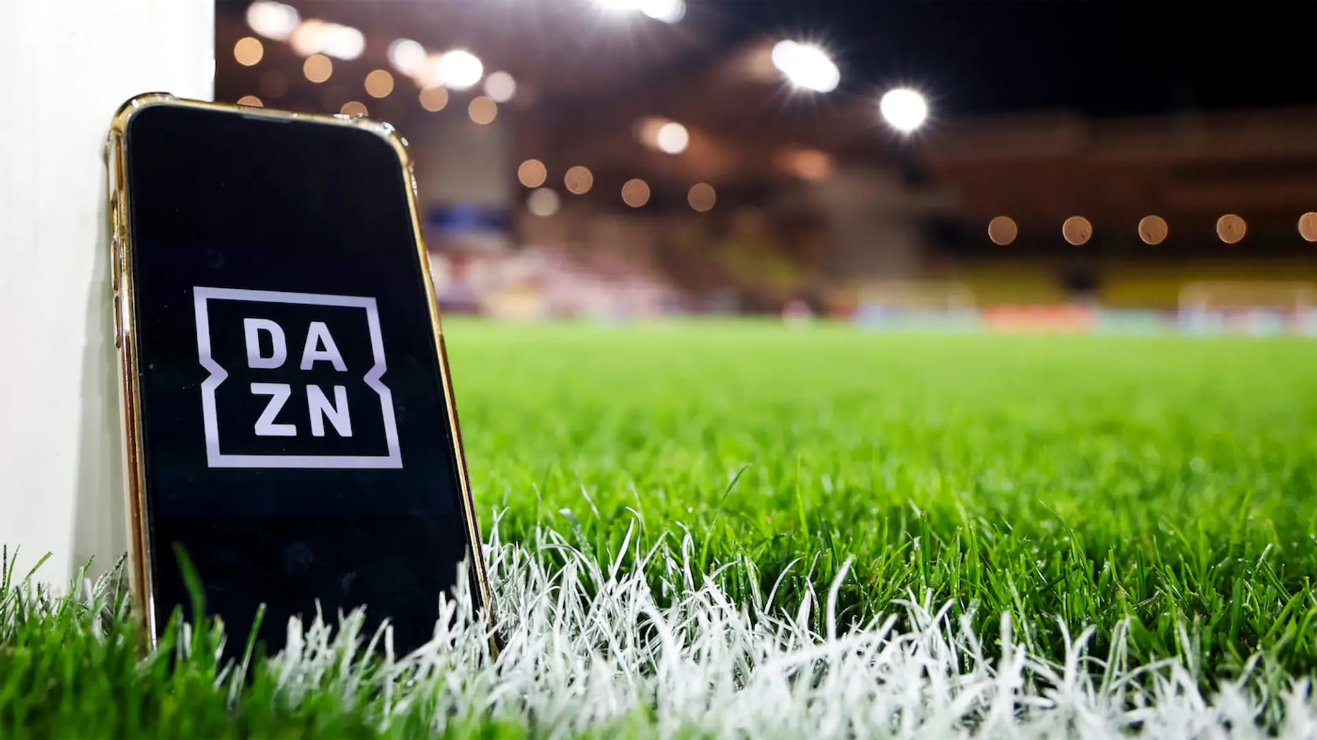 Smartphone con el logo de DAZN apoyado en el poste de la portería de un estadio de fútbol de noche.