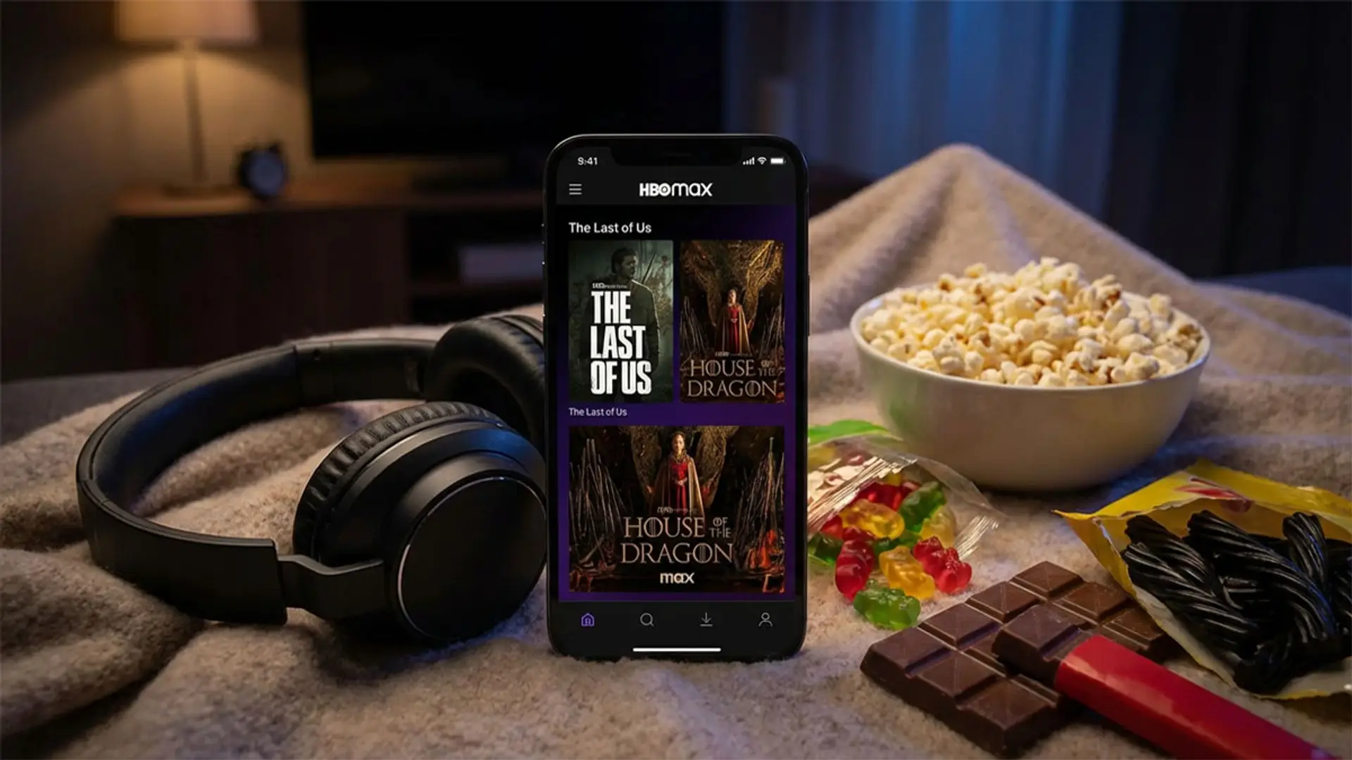 Smartphone con la interfaz de HBO Max mostrando series, junto a auriculares, palomitas y snacks en un entorno doméstico.