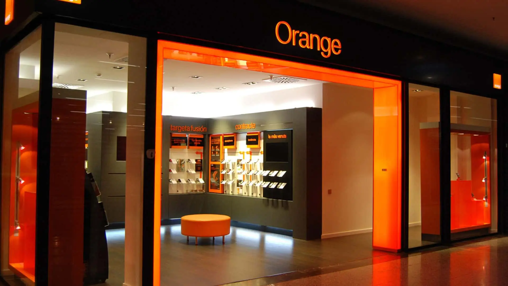 La venta de MasOrange avanza, pero 1.000 millones separan a Orange y los fondos dueños de MásMóvil Interior iluminado de una tienda Orange con expositores de teléfonos y zona de atención al cliente.