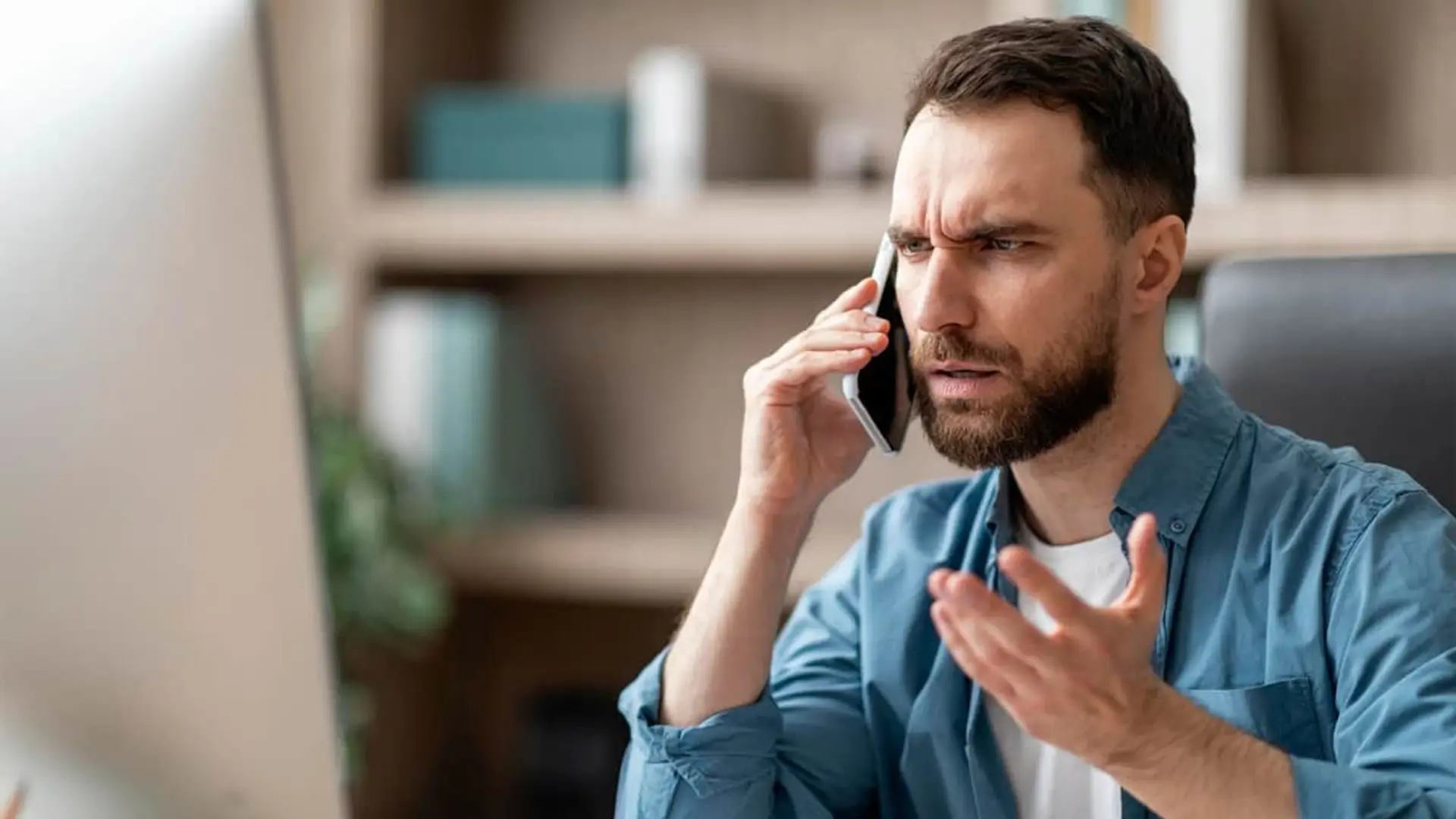 Hombre con expresión de enfado hablando por teléfono frente al ordenador para reclamar una estafa o incidencia.