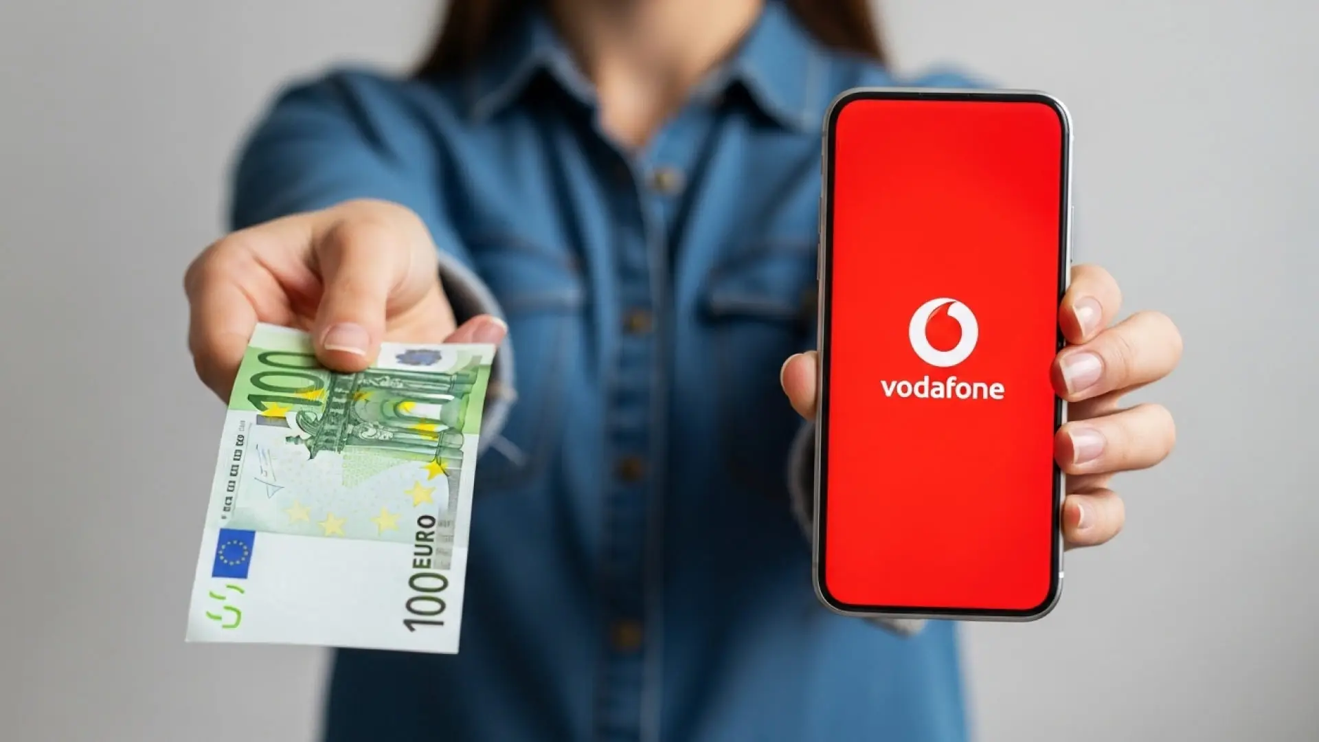 Vodafone te regala 100€ en metálico si haces esto Persona mostrando un billete de 100 euros y un móvil con el logo de Vodafone, por una promoción de la operadora.