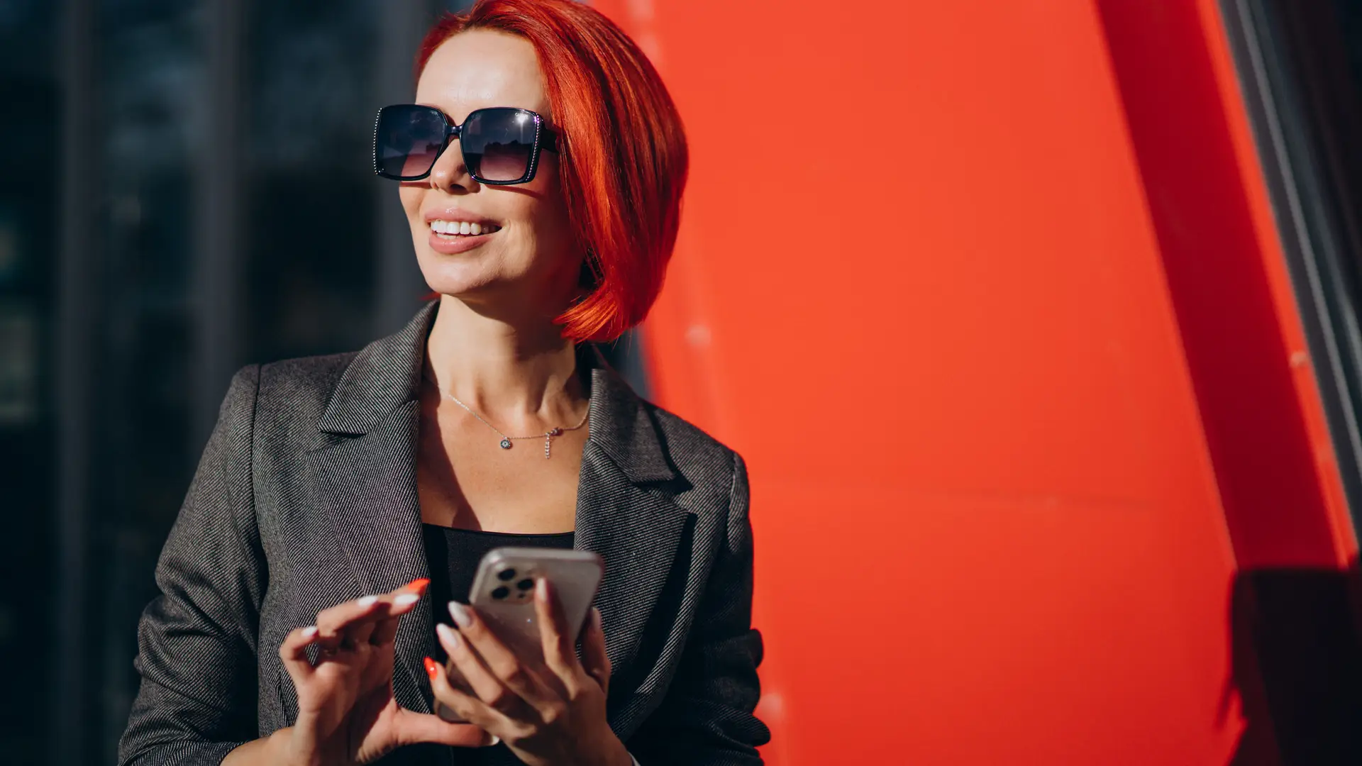 Mujer con gafas de sol y pelo rojo sonrie mientras utiliza su teléfono con tarifa cablemovil