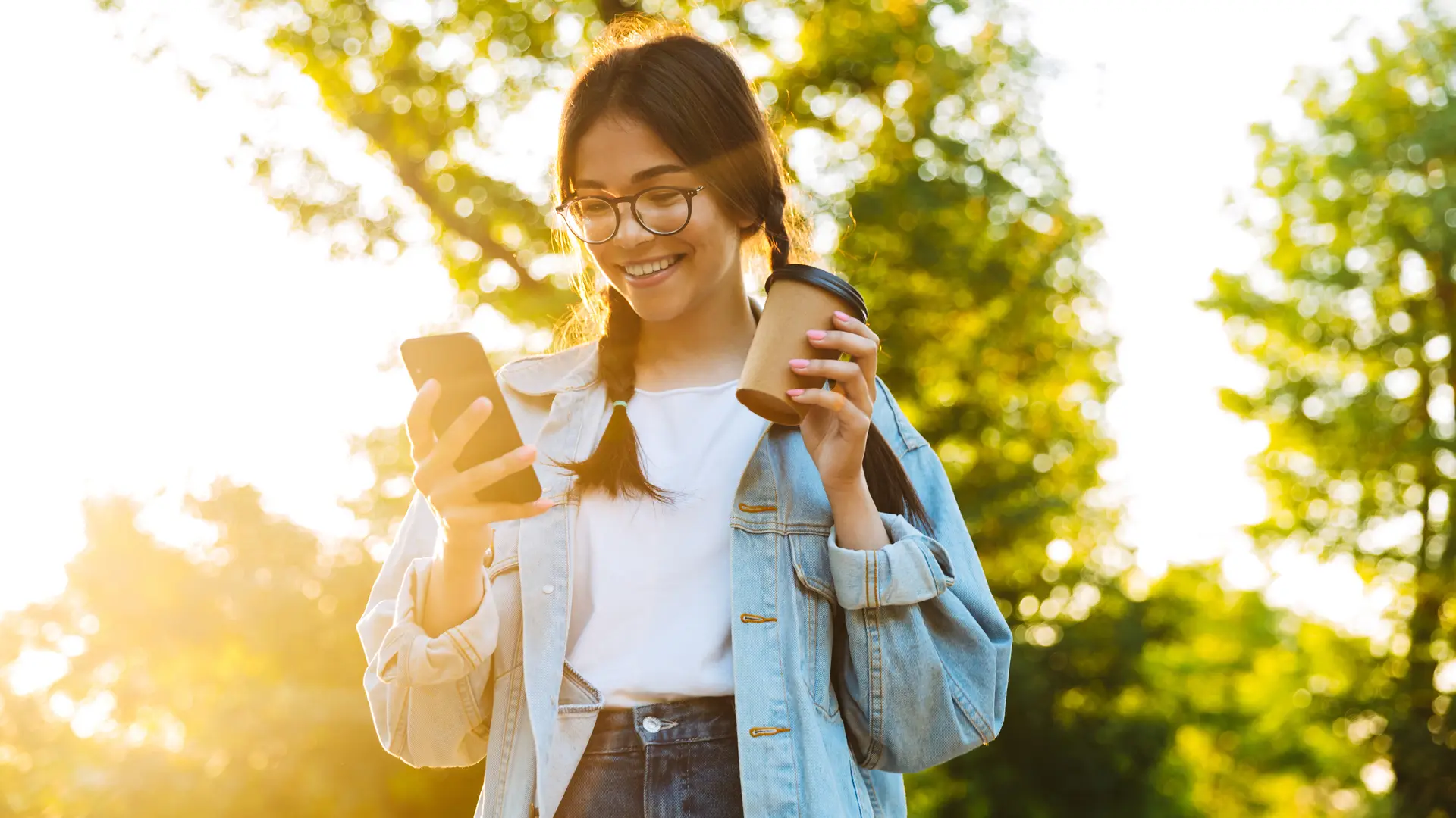 Joven sonrie en el parque y sujeta un café, mientras mira en su móvil las tarifas de excom Joven sonrie en el parque y sujeta un café, mientras mira en su móvil las tarifas de excom
