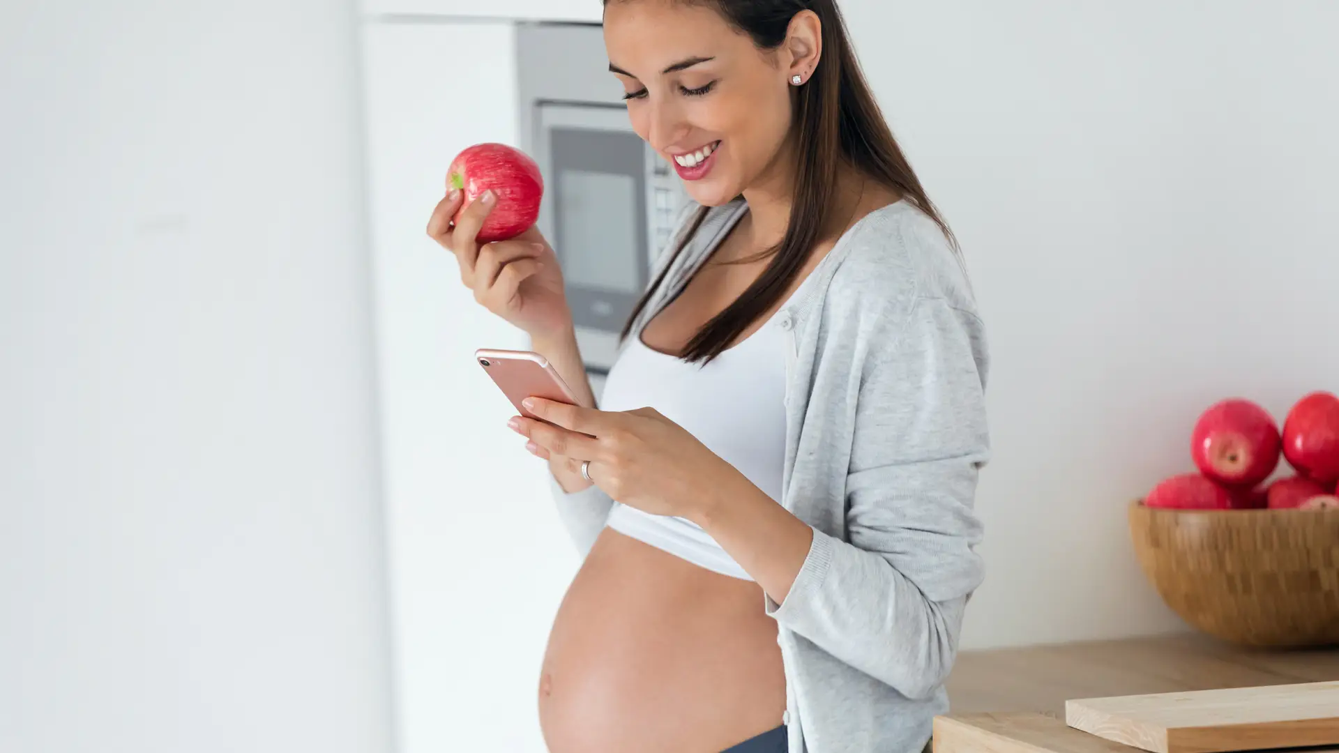 Mujer embarazada usando su smartphone mientras come una manzana simboliza línea adicional finetwork Mujer embarazada usando su smartphone mientras come una manzana simboliza línea adicional finetwork
