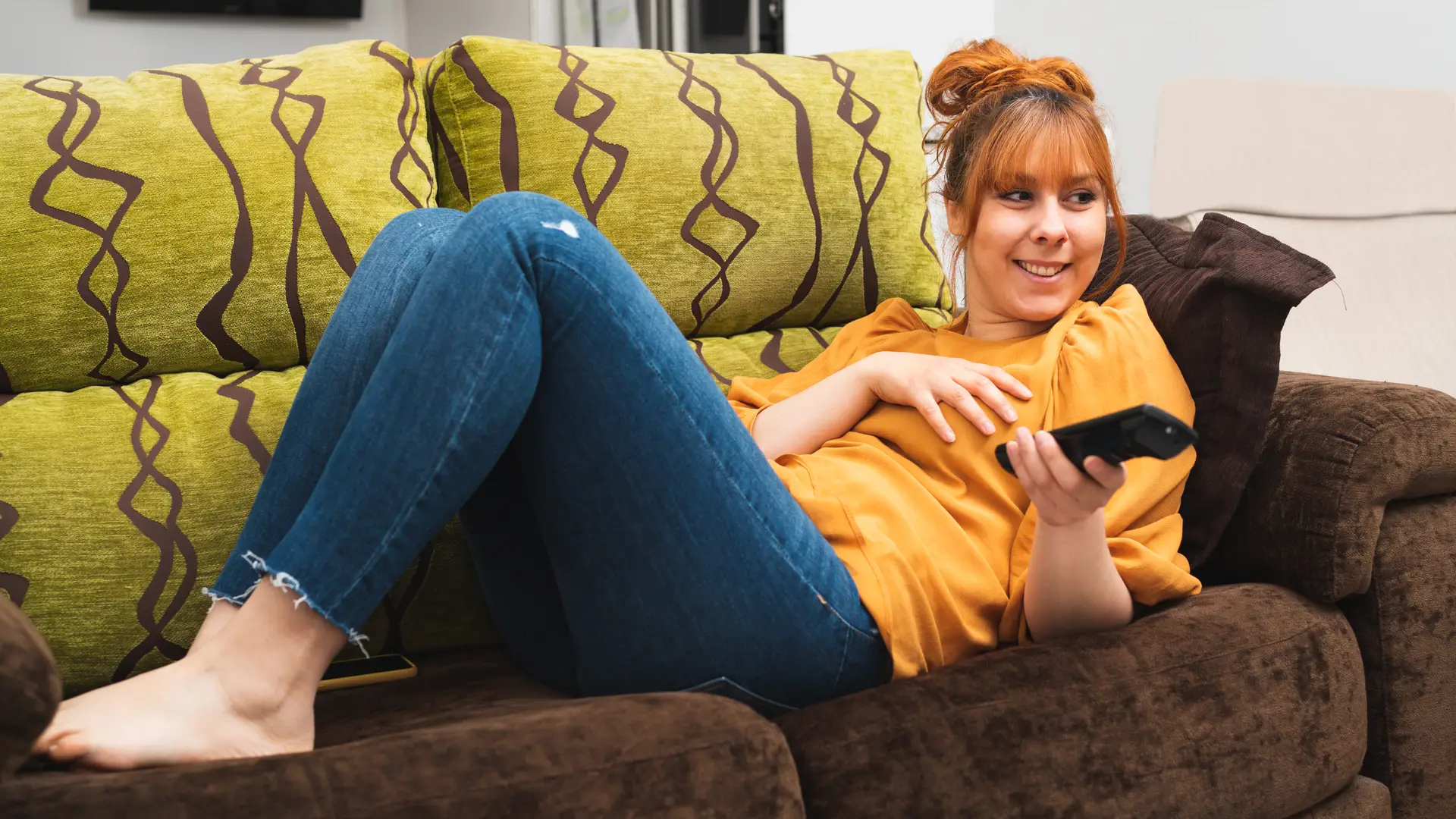 Mujer con camiseta amarilla ve el televisor desde su sofá mientras cambia de canal Mujer con camiseta amarilla ve el televisor desde su sofá mientras cambia de canal