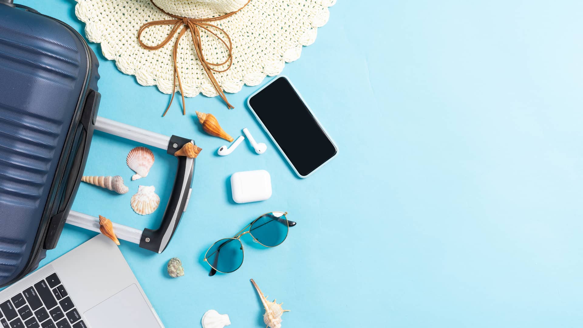 Móvil, maleta, portátil, gafas de sol, sombrero y conchas sobre suelo azul para hablar de los bonos extra de o2, ideales para aquellos de vacaciones
