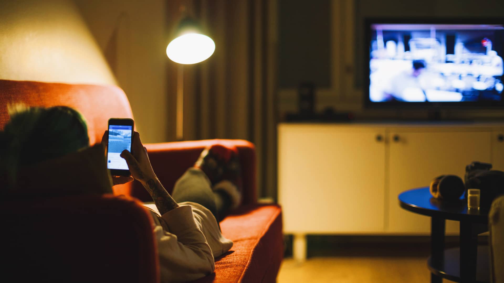 Persona tumbada en sofá navega por redes sociales en su móvil mientras ve la televisión, disfrutando de una tarifa de móvil y tv de orange