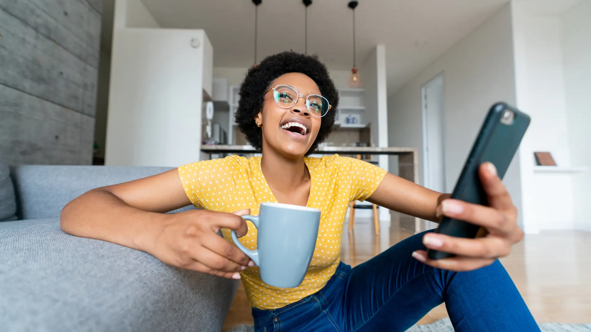 Mujer sentada en el suelo sonríe feliz al escuchar los mensajes de su buzón de voz de su móvil con tarifa de parlem mientras toma un café