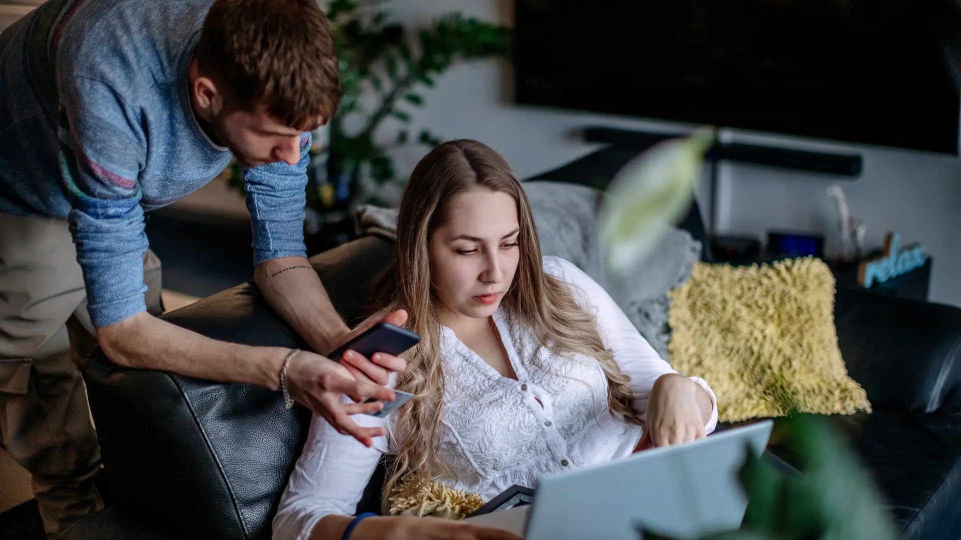 Pareja hace gestiones en su móvil y ordenador portátil desde el sofá con la tele de fondo, disfrutan de una tarifa de móvil, internet y televisión de pepephone Pareja hace gestiones en su móvil y ordenador portátil desde el sofá con la tele de fondo, disfrutan de una tarifa de móvil, internet y televisión de pepephone
