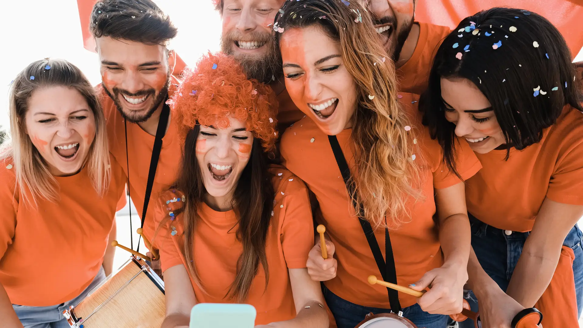 Grupo de amigos vestidos de naranja siguen un partido de su equipo favorito en el teléfono de una amiga porque aun no han contratado líneas adicionales con simyo